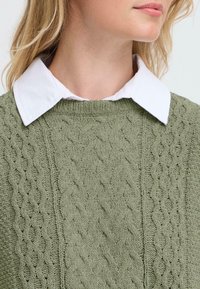 Olijfgroene kabelgebreide sweater met een ribgebreide ronde halslijn en een gelaagde witte coltrui eronder. Textuurpatroon over de hele sweater.