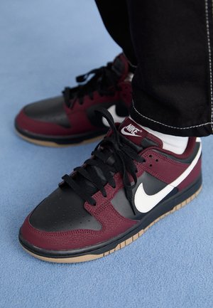 Personne portant des baskets Nike noires et bordeaux avec un logo swoosh blanc et des lacets noirs, debout sur un sol recouvert de moquette bleue.