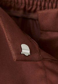 Petite épingle argentée en forme de cloche attachée au col d'un vêtement en tissu marron texturé.