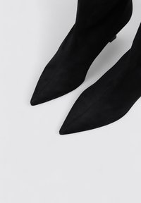 Botas de tobillo de ante negro con puntera afilada y tacón delgado. Presentan una textura suave y un diseño elegante, con un detalle de costura mínimo.