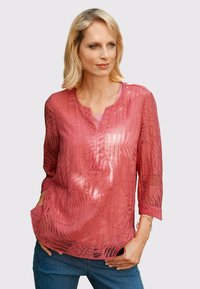 Transparente, leichte rote Bluse mit Blattmuster, Dreiviertelärmeln, V-Ausschnitt und Vorderentaschen; über einem hellen Oberteil getragen.