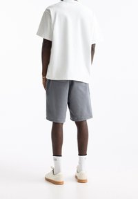 Biała bawełniana koszulka i szare szorty, prezentujące luźny krój. Białe trampki z gumową podeszwą i widoczne skarpetki do kostek. Całość w prostym designie.