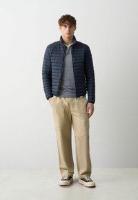 Veste doudoune navy sur un pull gris, associée à un pantalon cargo beige et des baskets marron. Comprend une fermeture éclair et plusieurs poches.