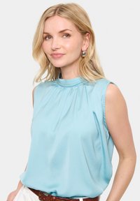 Blusa senza maniche di un azzurro chiaro con collo alto, dettagli arricciati e texture liscia, abbinata a una cintura marrone.