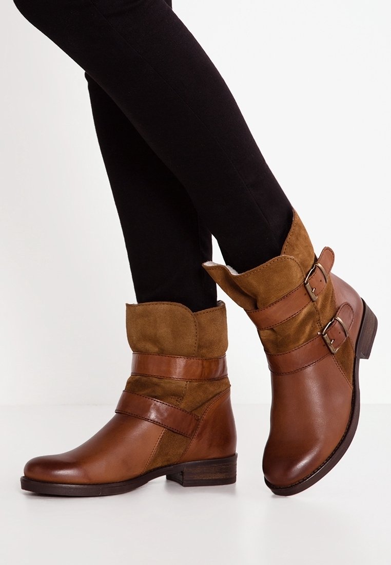 Bottines en cuir marron et suède avec double bride à boucle portées avec un pantalon noir sur fond blanc.