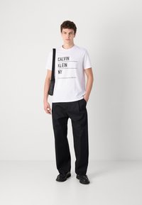 Bijela pamučna majica s crnim natpisom "CALVIN KLEIN NY" i logom, uparena s crnim širokim hlačama i crnim cipelama. Muškarac drži torbu.