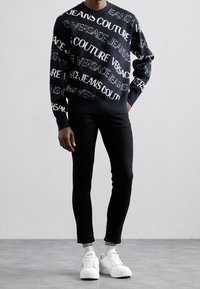 Suéter de punto negro con estampado del logo en blanco "Versace Jeans Couture", combinado con pantalones negros de corte slim y zapatillas blancas con calcetines a rayas.