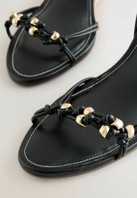 Sandales plates noires avec fines brides nouées et accents de perles dorées, avec surpiqûres blanches autour de la zone ovale des orteils.