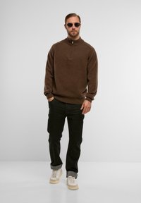 Brauner Strickpullover mit hohem Kragen und Reißverschluss, kombiniert mit schwarzen Jeans und weißen Sneakern mit einer cremefarbenen Sohle. Sonnenbrille rundet das Outfit ab.