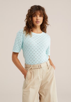 Lokkis juustega naine, kes kannab helesinist lühikeste varrukatega kootud pluusi ja beeži värvliga kõrge vöökohaga pükse, käed taskutes, neutraalne taust.