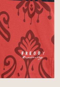 Tessuto rosso con un motivo marrone che si avvolge. Nell'angolo inferiore è presente la scritta "FREDDY" in grassetto bianco e "training" in corsivo.