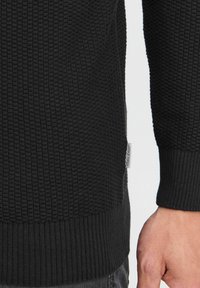 Pull en tricot noir avec une finition texturée, des bords-côtes aux poignets et à l'ourlet. Présente une petite étiquette sur le côté.