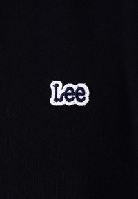 Svart stoff med en liten hvit og mørkeblå brodert "Lee"-logo i midten.