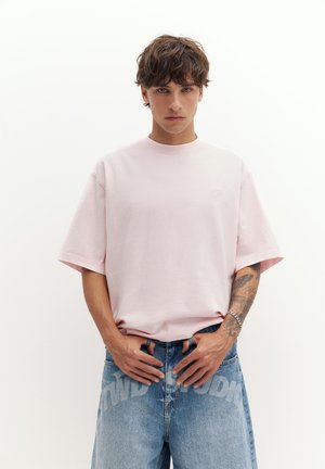 Jeune homme aux cheveux bruns bouclés portant un t-shirt oversize rose clair et un jean en denim bleu avec un texte graphique, debout devant un fond blanc uni.