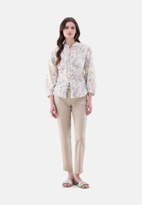 Floral-patterned blouse in white, yellow, and gray, featuring ruffles and a mandarin collar, paired with beige fitted pants and sandals.
