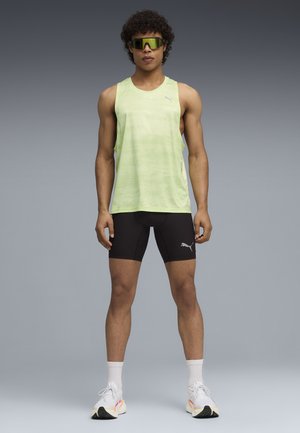 Lichtgroen, textuurvol singlet gecombineerd met zwarte, nauwsluitende short met een logo. Model draagt grote groene zonnebril en hardloopschoenen.