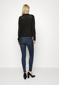 Mujer con cabello largo rubio que lleva una blusa de encaje negro de manga larga, jeans ajustados azul oscuro y zapatos de tacón negro.
