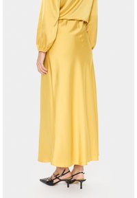 Gonna maxi satin gialla con un design fluido, dotata di una vita arricciata e una texture liscia, abbinata a tacchi neri a punta.