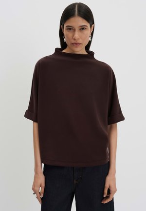 Vrouw met steil donker haar draagt een losse donkerbruine korte mouw top met opgerolde boorden en donkere spijkerbroek, kijkt vooruit tegen een effen achtergrond.