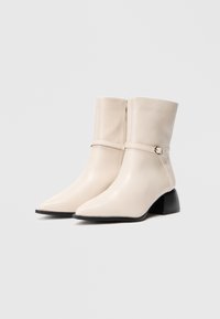 Par af beige spidse ankelstøvler med lave chunky sorte hæle og sideskilt med dekorrativ rem og spænde.