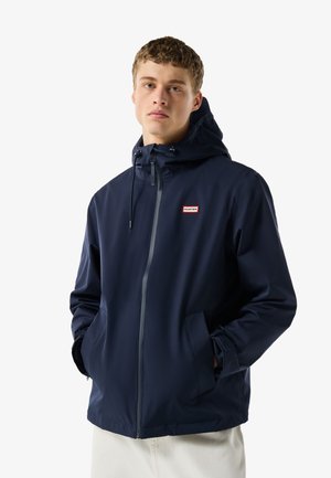 Mladenič v mornarsko modri kapucarji z zadrgo spredaj in rokami v žepih, stoji pred belo ozadje.