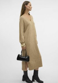 Robe en maille beige avec un col en V, des manches longues, une fente sur le côté, associée à des bottines noires à talons et un petit sac à main noir.