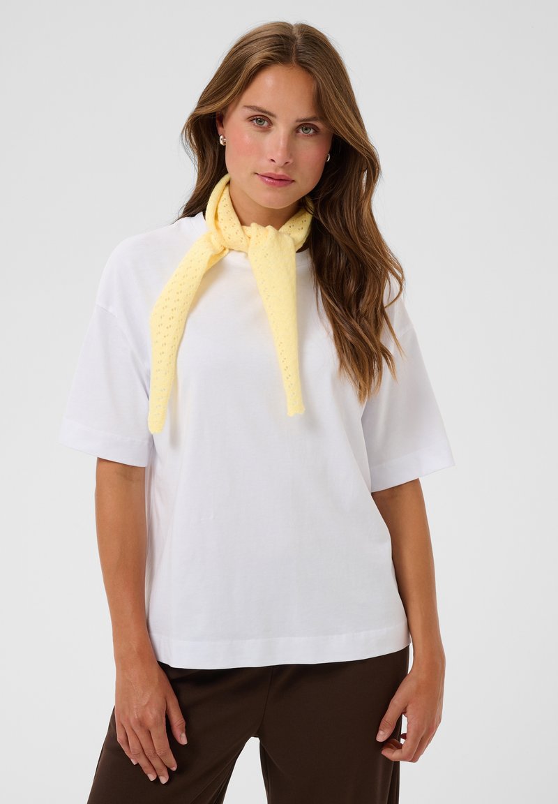 Femme aux longs cheveux bruns portant une chemise blanche à manches courtes, un pantalon marron et une écharpe jaune nouée autour du cou sur un fond gris.
