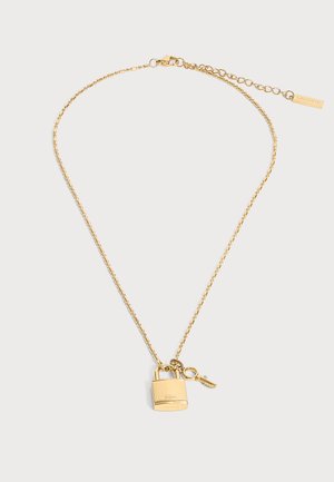 Guldhalskæde med en lille hængelås og nøglevedhæng, med Lacoste-logo og justerbar lås med mærkeetiket.