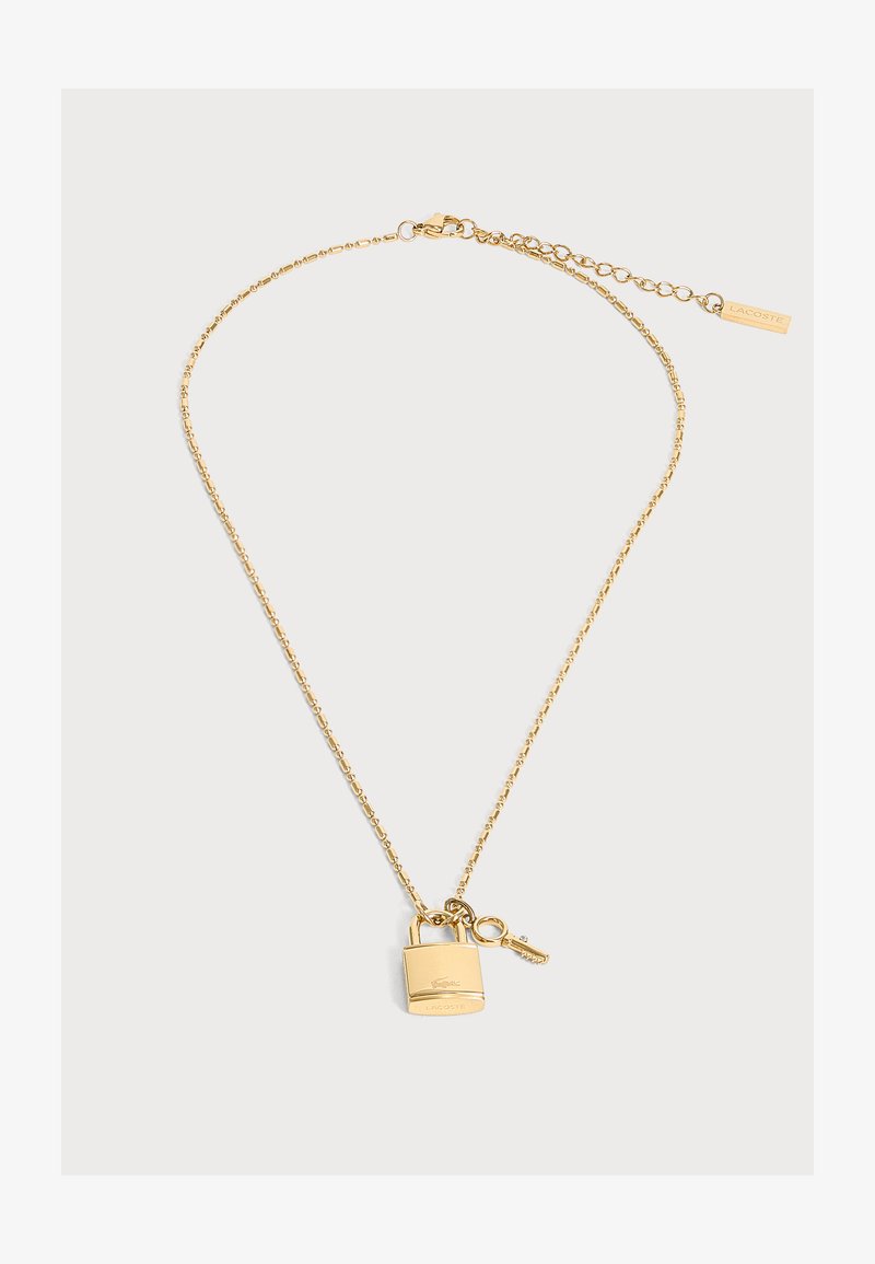 Collier chaîne en or avec petit cadenas et pendentif clé, avec logo Lacoste et fermoir ajustable avec étiquette de marque.