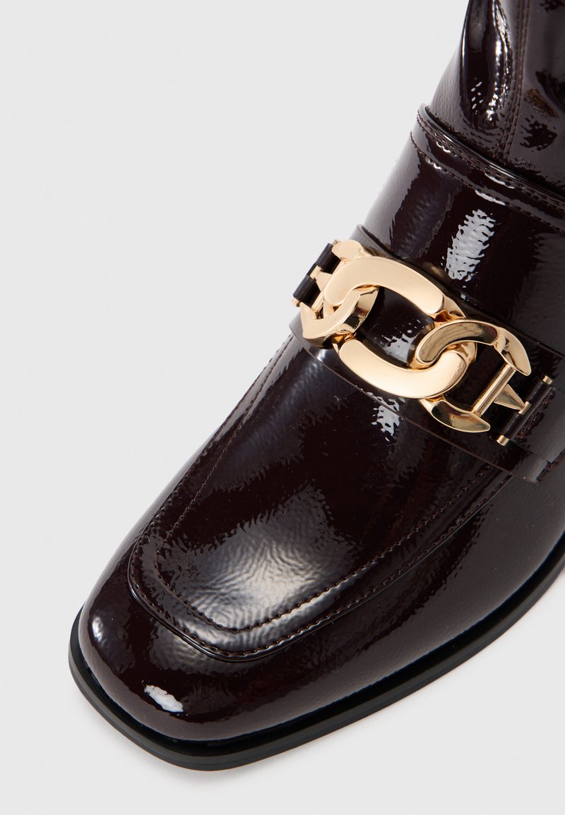 Scarpa in pelle verniciata marrone scuro con punta quadrata, caratterizzata da un accessorio a catena dorata lungo il cinturino alla caviglia e una superficie liscia e lucida.