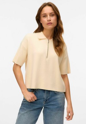 Hellbeige Poloshirt mit kurzen Ärmeln und einem halben Reißverschluss, aus strukturiertem Stoff gefertigt. Kombiniert mit hochgeschnittenen blauen Jeans.