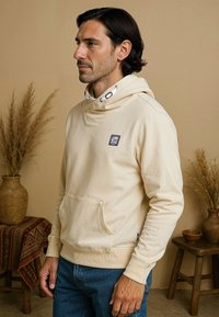 Hoodie beige avec une poche avant, doté d'une capuche intérieure à motifs et d'un patch logo sur la poitrine. Texture de tissu douce.