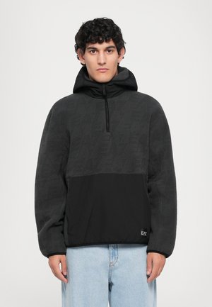 Mann som har på seg en svart og mørkegrå hettegenser med frontglidelås og lyseblå jeans, vendt fremover mot en hvit bakgrunn.