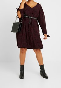 Schwarz-burgunderrotes Kleid mit Animal-Print, V-Ausschnitt, langen Ärmeln und Schlaufen-Details, tailliert mit einem Nieten-Gürtel. Kombiniert mit schwarzen Stiefeletten und einer quadratischen Tasche.