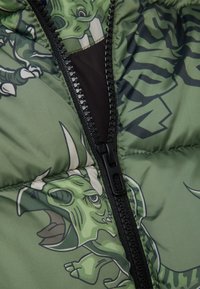 Chaqueta acolchada verde con cremallera y patrón de dinosaurios de dibujos animados. El material es suave, con un acabado brillante y un forro interior suave.