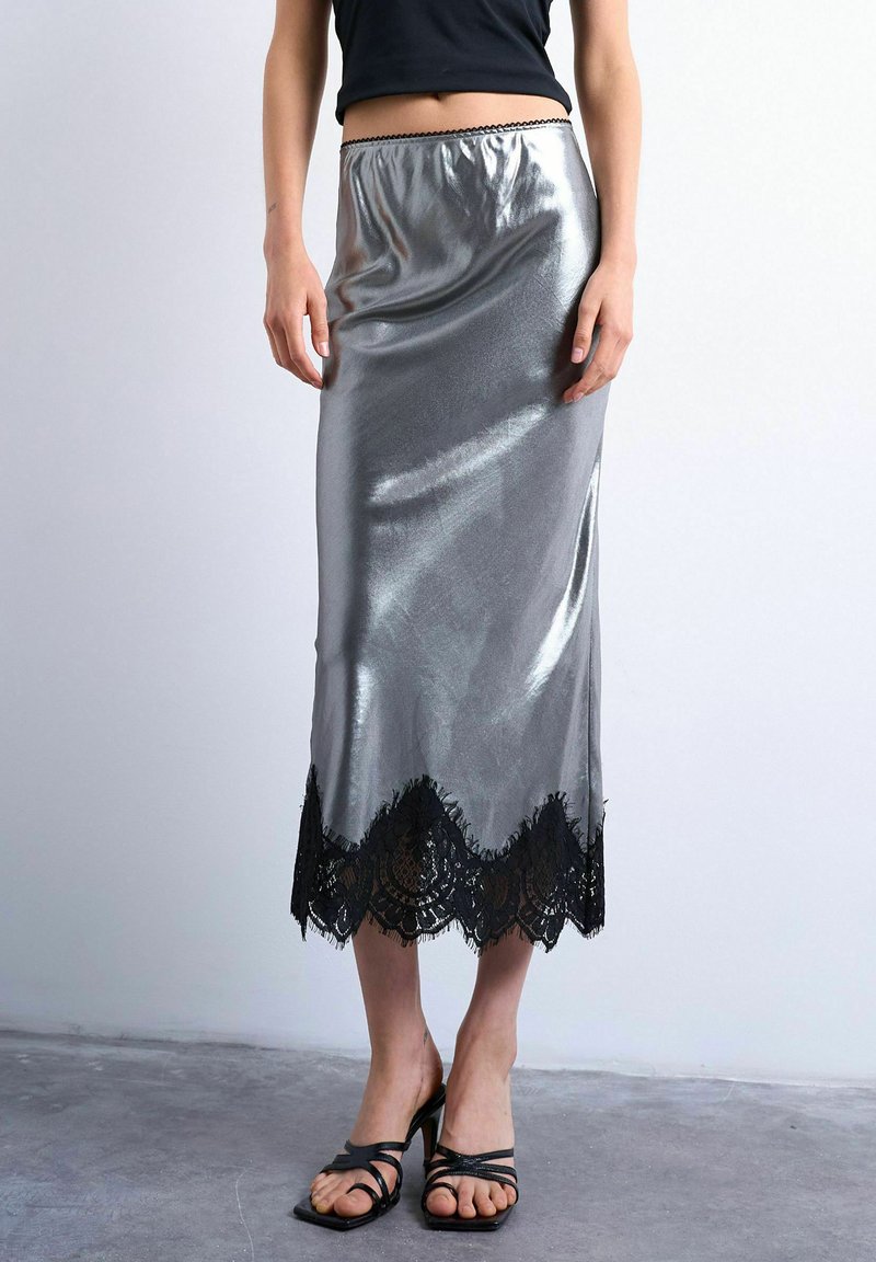 Personne portant une jupe midi en satin argenté avec un bord en dentelle noire et des sandales à talons hauts noires à lanières, debout sur un sol en béton.