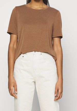 Person trägt ein braunes T-Shirt mit kurzen Ärmeln, leicht in hoch taillierte, cremefarbene Hosen mit sichtbaren Knöpfen und Taschen gesteckt.