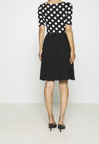 Musta mekko, jossa on vartalonmyötäinen yläosa valkoisilla pilkuilla, lyhyet hihat ja liehuva helma; yhdistetty avoimilla mustilla avokkailla.