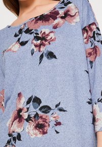 Blus med blått blommönster som har en rundad halsringning, trekvartslånga ärmar och ett mjukt tyg. Blommönstret inkluderar rosa och vinröda blommor med mörka blad.