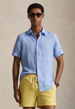 Homme portant des lunettes de soleil, une chemise à manches courtes bleu clair boutonnée, un short jaune à cordon de serrage et une montre-bracelet sur fond neutre.