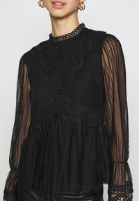 Top nero con un corpino in pizzo dalle piccole arricciature, maniche plissettate trasparenti e un collo alto con dettagli di rifinitura intricati.