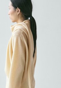 Beżowy sweter dziergany z luźnym kapturem, ukazujący miękką fakturę i opadające ramiona, widziany z boku na jasnym tle.