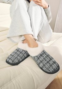 Black and white woven slippers with soft white fur trim, rubber soles, and a rounded toe design, resting on a textured beige surface.