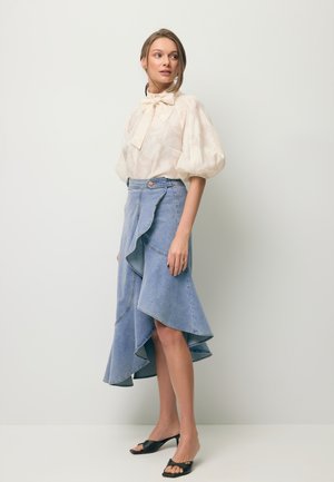 Vrouw draagt een blouse met pofmouwen en een strik, en een asymmetrische gerimpelde denim midi-rok met zwarte open-teen hakken.