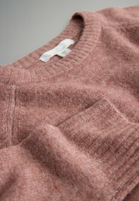 Różowy sweter z dzianiny z prążkowanym dekoltem i hemem, z kieszonką z przodu. Materiał wydaje się miękki o teksturowanej powierzchni. Widoczna metka z zaleceniami dotyczącymi pielęgnacji.