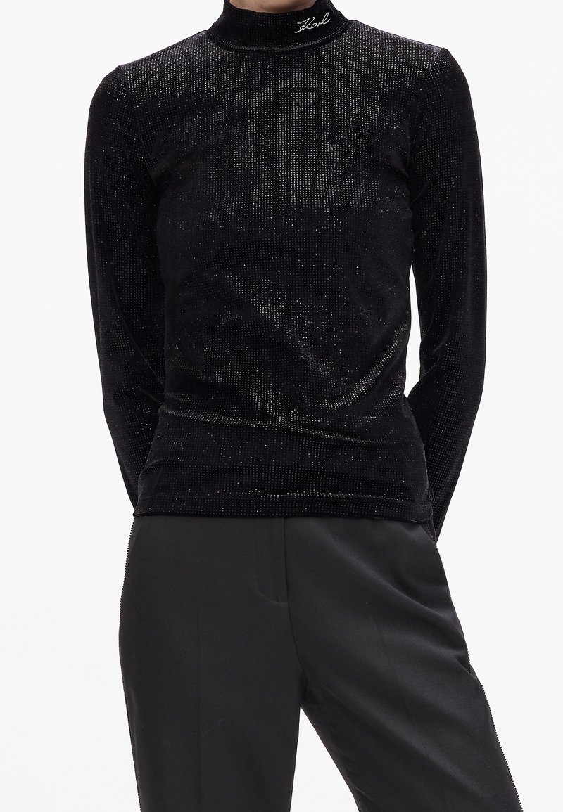 Personne portant un haut noir moulant à manches longues avec une texture subtile scintillante et un pantalon noir sur fond blanc.