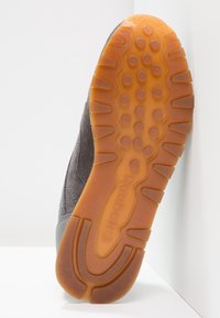 Baskets en suède gris avec une semelle texturée, présentant une semelle en caoutchouc marron et des motifs de grip circulaires. Logo visible sur le côté.