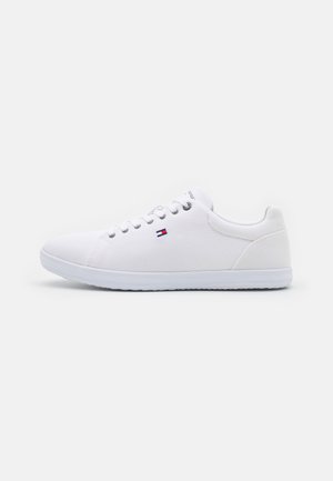 Zapatilla blanca de tipo bajo con parte superior de tela, suela blanca, ojales metálicos y pequeño logo rojo, blanco y azul en el lateral.
