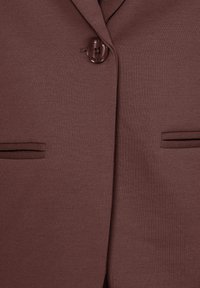 Gros plan de l'avant d'un blazer marron avec un seul bouton foncé et deux poches fendues horizontales de chaque côté.