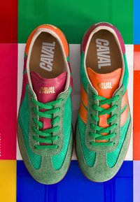 Paire de baskets en daim vert avec lacets verts, accents en cuir orange et grenat, marquage « CAVAL » à l'intérieur et sur les languettes, sur un sol en carreaux colorés.